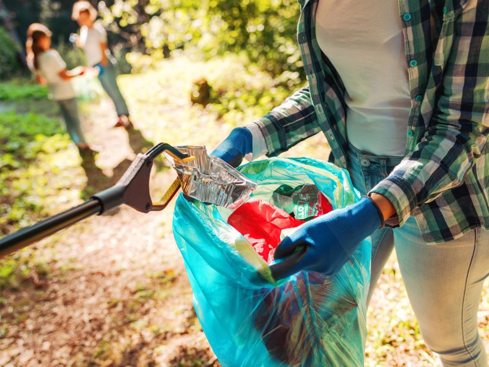 Eine Frau hält eine Mülltüte in der Hand, in die per Greifarm eine Aluminiumschale geworfen wird.____