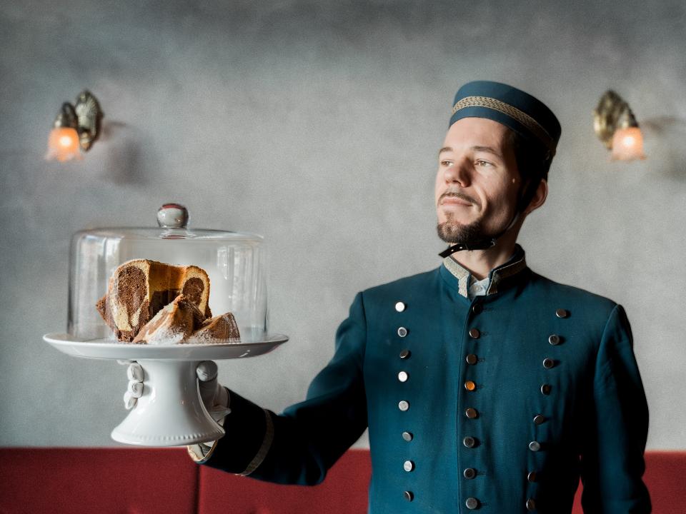 Kellner in blauer Uniform präsentiert Kuchen unter einer Glasglocke in einem stilvollen Café.____