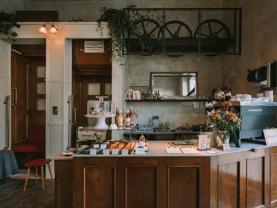 Holzgetäfelte Café-Theke mit frischen Blumen und Kuchen, im Hintergrund eine elegante Holztür.____
