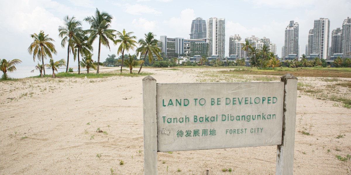 Schild mit Aufschrift "Land to be developed" an leerem Strand mit Stadt im Hintergrund