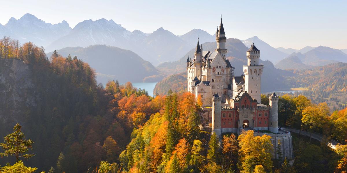 Schloss Neuschwanstein auf einem Hügel mit herbstlichen Bäumen vor See- und Bergkulisse.
