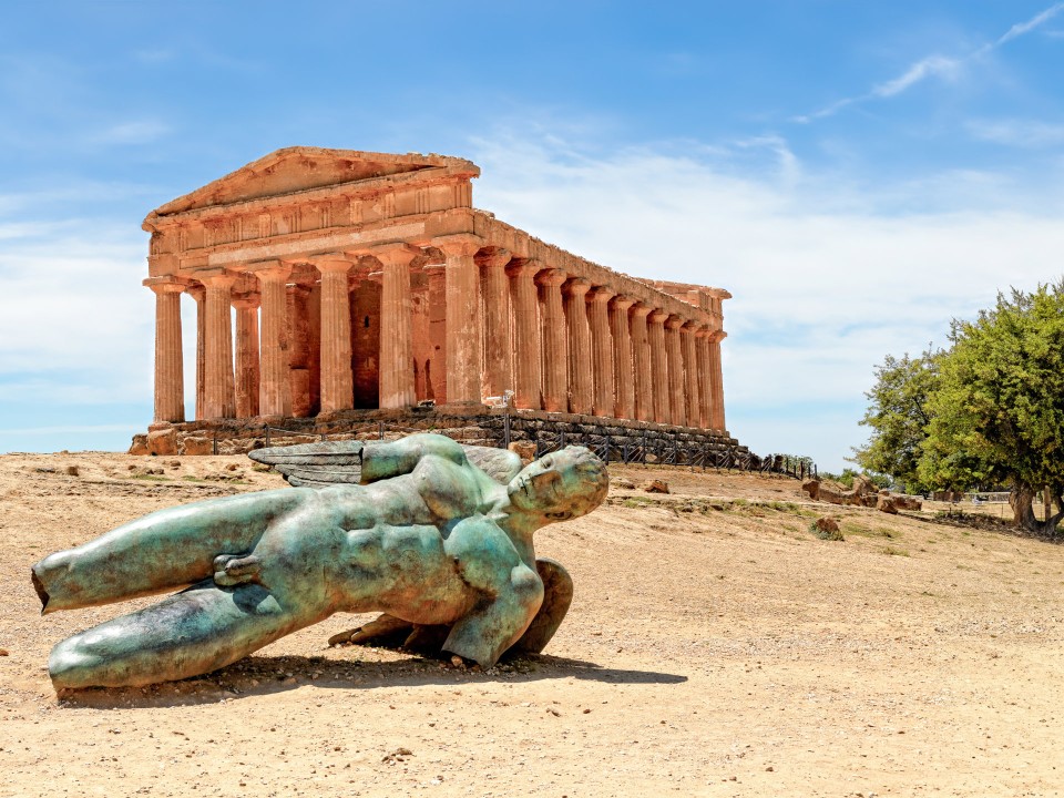 Eine Marmorstatue liegt auf dem Sandboden. Es ist die Statue "Gestürzter Ikarus" auf Sizilien.____