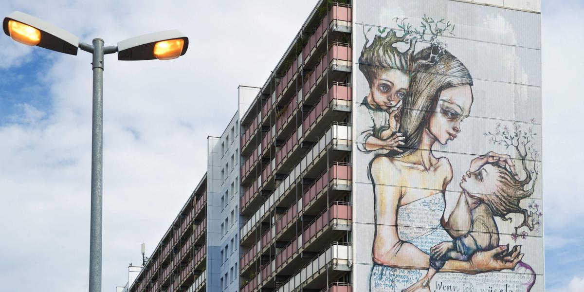 Hochhaus mit großem Wandgemälde einer Frau und zweier Kinder. Laterne im Vordergrund, bewölkter Himmel im Hintergrund.