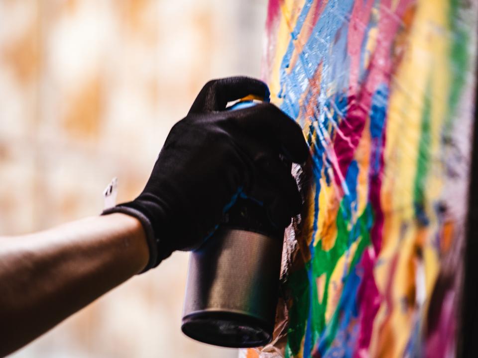 Nahaufnahme einer Hand mit schwarzem Handschuh, die eine Sprühdose hält, vor einer bunten Graffiti-Wand.____