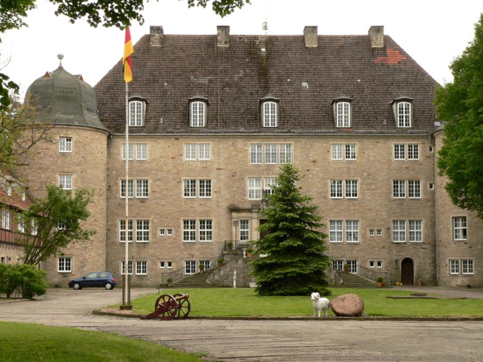 Schloss in bräunlichen Farben vor einem Innenhof mit Rasenflächen und Baum in der Mitte.____