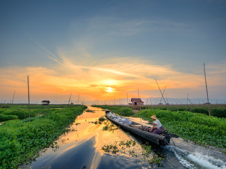 Eine Person fährt auf einem Boot in Richtung Sonnenuntergang, links und rechts sind schwimmende Bete auf dem Wasser.____