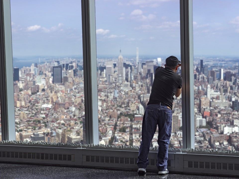 Ein Mann fotografiert durch eine große Fensterfront hindurch die Stadt New York.____