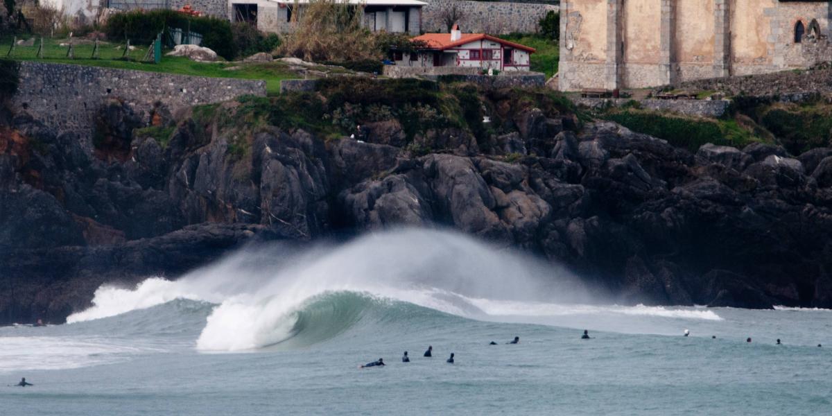Mehrere Surfer warten im Wasser vor steilen Felsen an einer alten Steinkirche hoch über der Küste