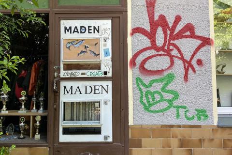 Automat mit der Aufschrift "Maden" und einem Fensterausschnitt, der Fische zeigt, an einer Graffiti-beschmierten Wand