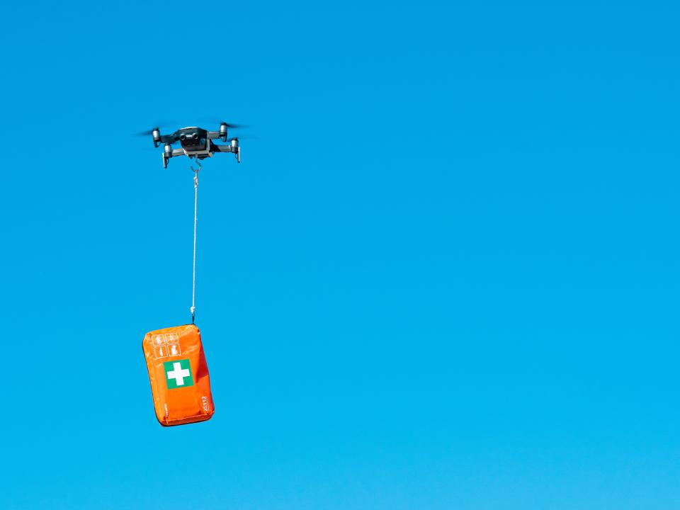Drohne transportiert eine orangefarbene Erste-Hilfe-Tasche am blauen Himmel____
