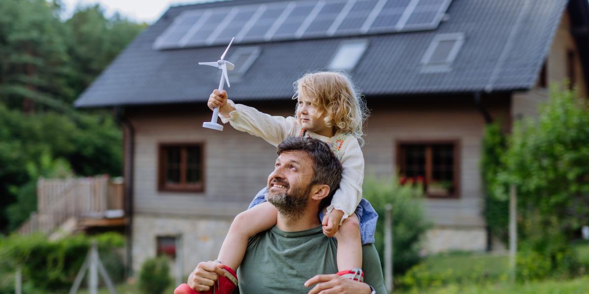 Glücklicher Vater trägt seine Tochter auf den Schultern vor einem Haus mit Solarpanelen. Das Mädchen hält eine kleine Windturbine