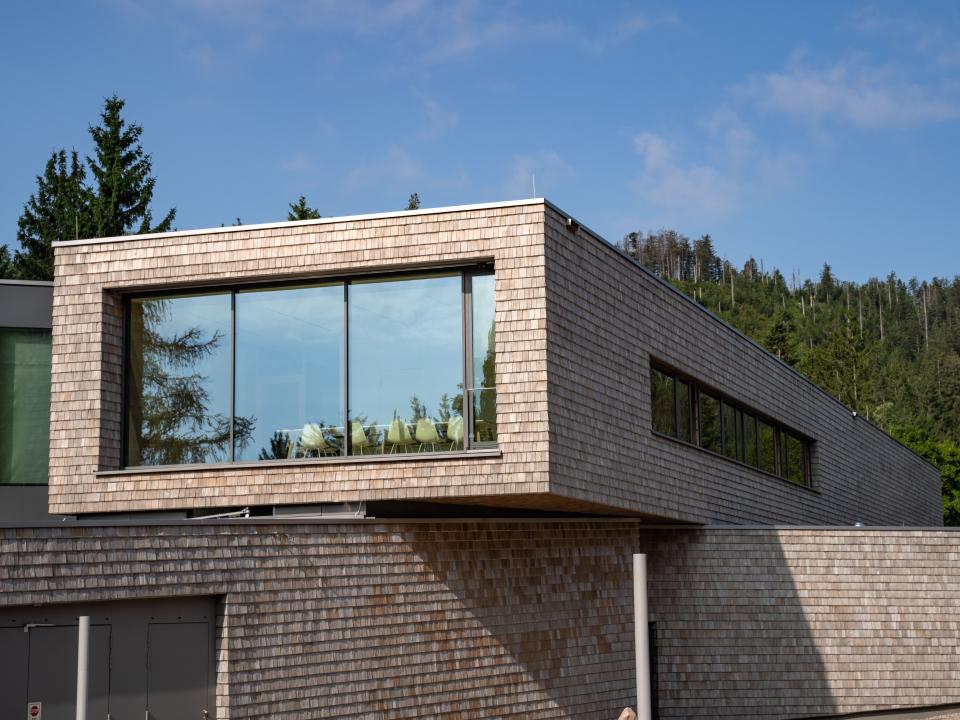 Moderne Architektur mit großen Fenstern, Holzverkleidung und Wald im Hintergrund bei sonnigem Wetter.____