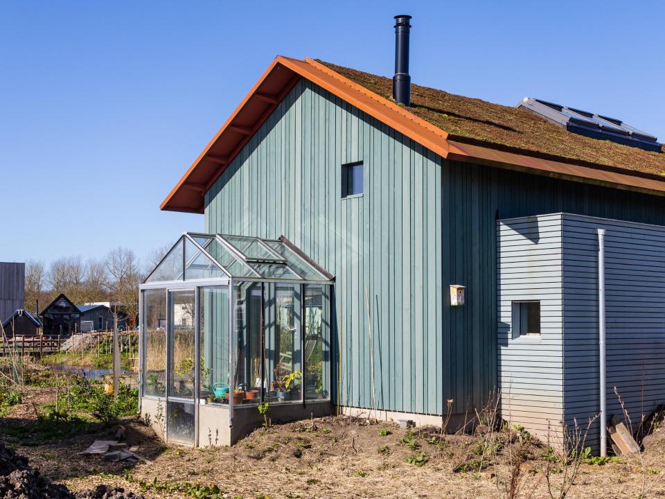 Holzhaus mit geneigtem, begrüntem Dach zeigt an der Seite einen verglasten Gewächshausanbau, in dem Pflanzgefäße stehen____