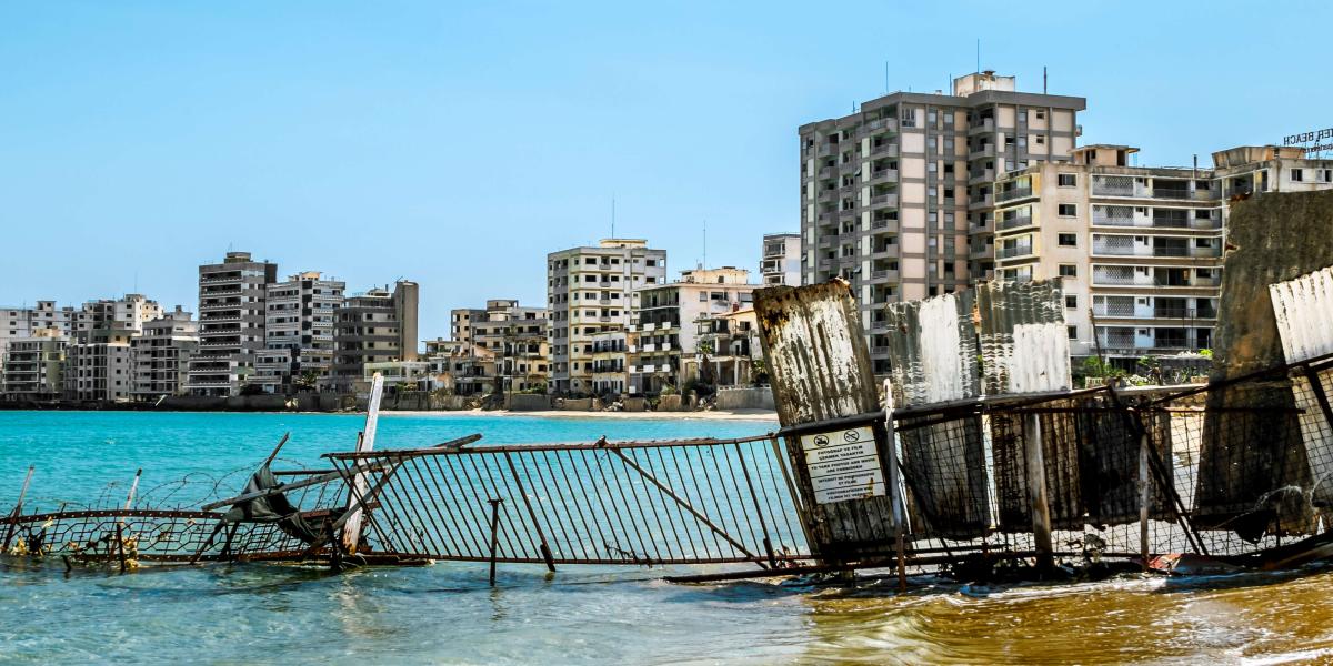 eilweise im türkisfarbenen Meer versunkener Zaun, dahinter eine Reihe verfallener Küstengebäude