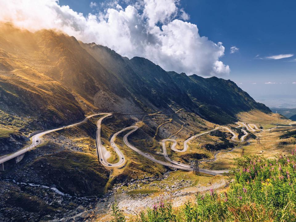 Serpentinenstraße schlängelt sich durch sonnige Berglandschaft mit steilen Hängen und dramatischem Himmel____
