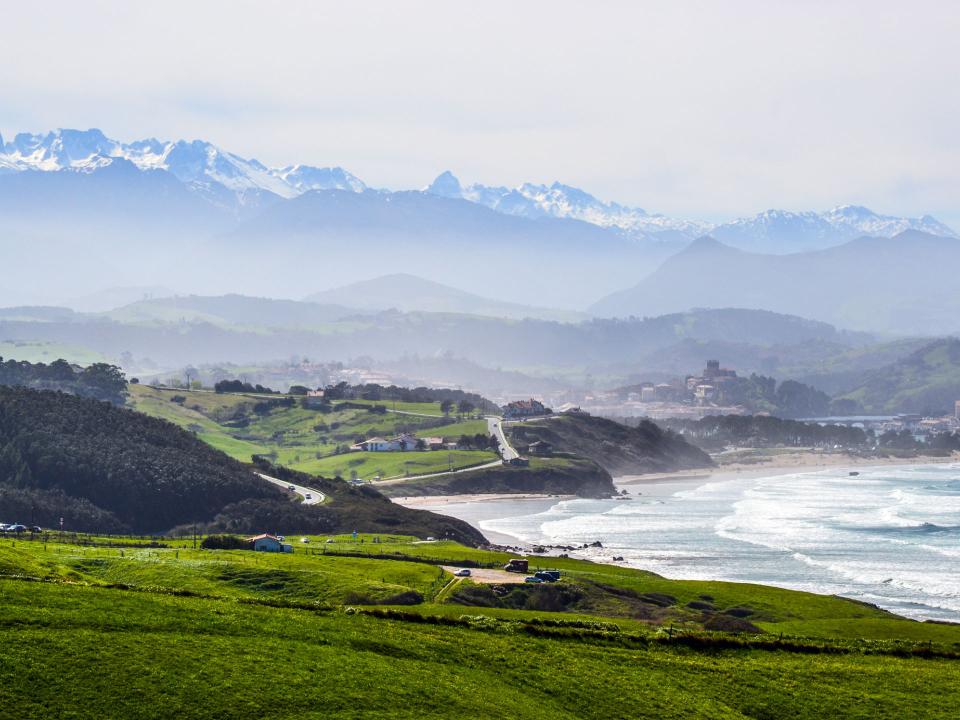 Grüne Hügel und Küstenstraße mit Blick auf schneebedeckte Berge und sanfte Meereswellen im Hintergrund____