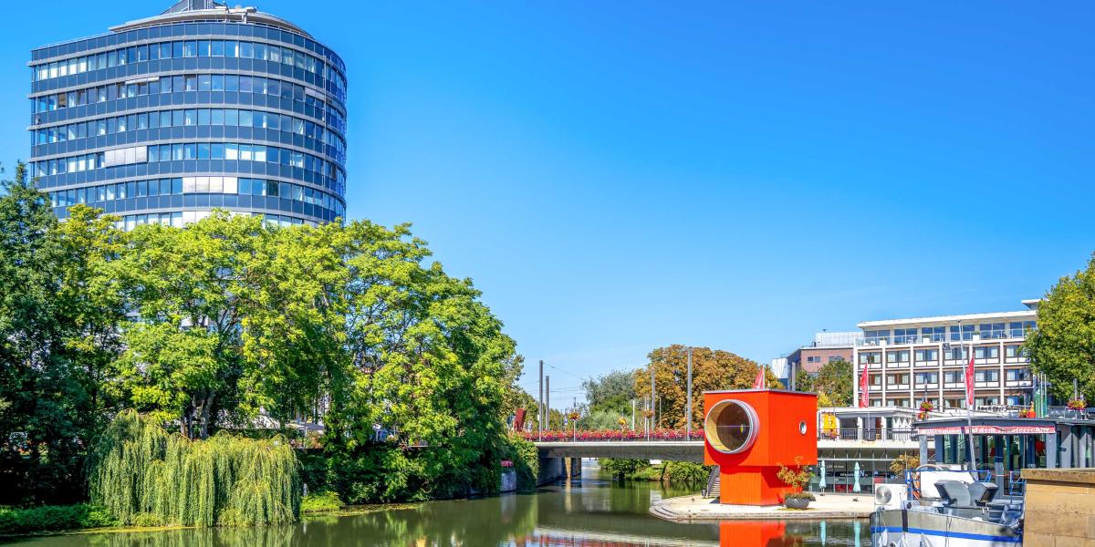 Idyllischer Flusslauf mit auffälligem rotem Kunstobjekt, einer Brücke und modernem, runden Hochhaus unter blauem Himmel