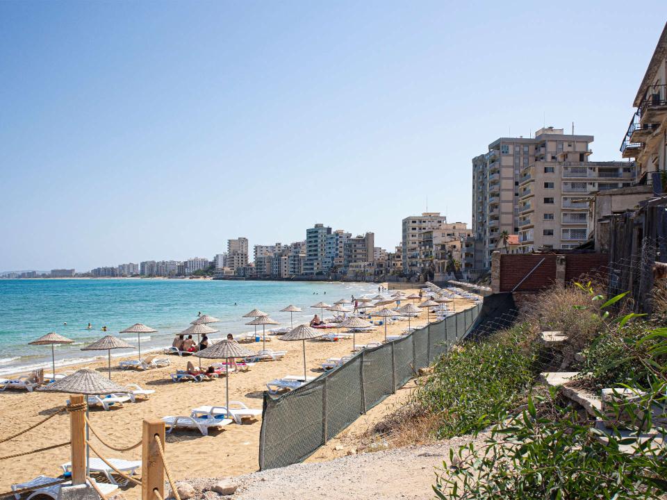 Türkisfarbenes Meer an belebtem Strand mit Sonnenschirmen, dahinter verlassene Hochhäuser____