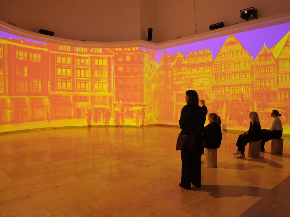 Besucherinnen betrachten leuchtend orange Projektion historischer Fassaden im Deutschen Pavillon, Biennale 2025____