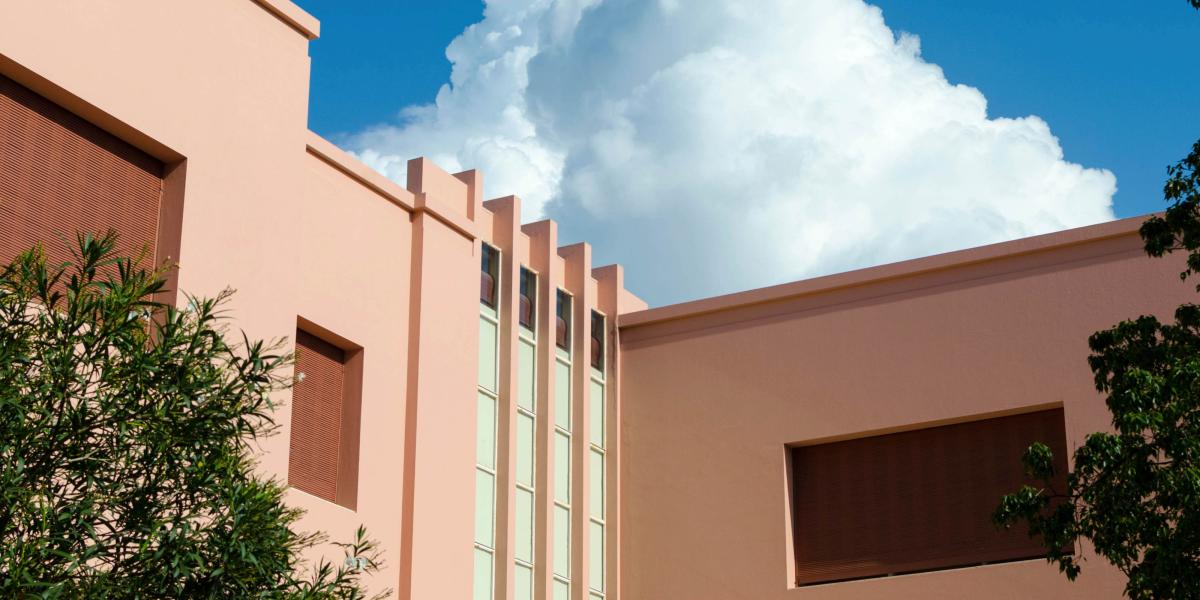 Modernes, pastellfarbenes Gebäude mit flachen Linien und großen Fenstern, umgeben von Bäumen, vor einem klaren blauen Himmel mit Wolken