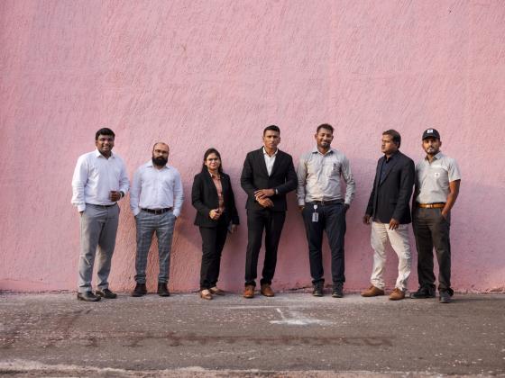 Gruppe von sieben Geschäftsleuten posiert vor einer rosa Wand für ein Gruppenfoto.