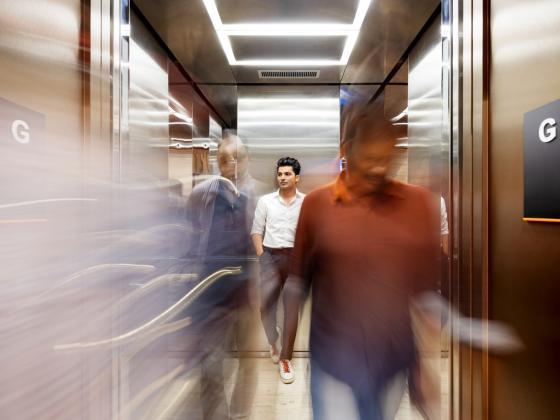 Menschen bewegen sich in einem modernen Aufzug, ein Mann steht ruhig in der Mitte.