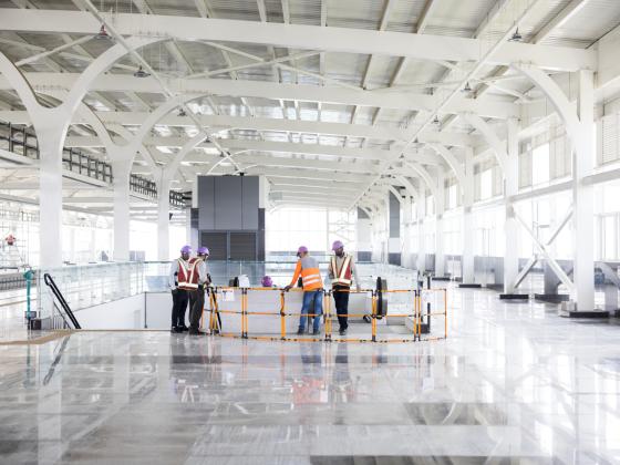 Bauarbeiter in Schutzhelmen und Westen besprechen in einer großen, leeren Halle.