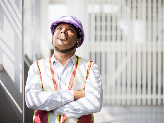 Bauleiter mit lila Schutzhelm und verschränkten Armen blickt nachdenklich nach oben.