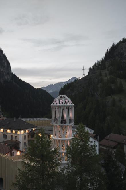 Beleuchteter Aussichtsturm mit spiralförmigen Säulen bei Dämmerung, umgeben von Bergen und traditionellen Häusern