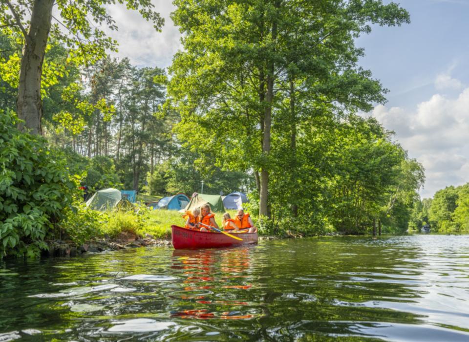 Kinder in roten Kanus mit Schwimmwesten vor bunten Zelten am grünen Ufer eines Waldbachs im Sommer.____