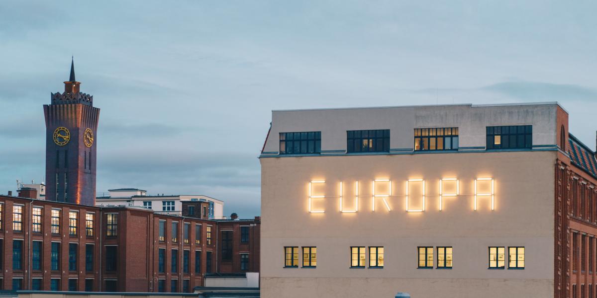 Fassade eines Gebäudes mit leuchtendem Schriftzug 'EUROPA', im Hintergrund ein historischer Turm mit goldenen Uhren