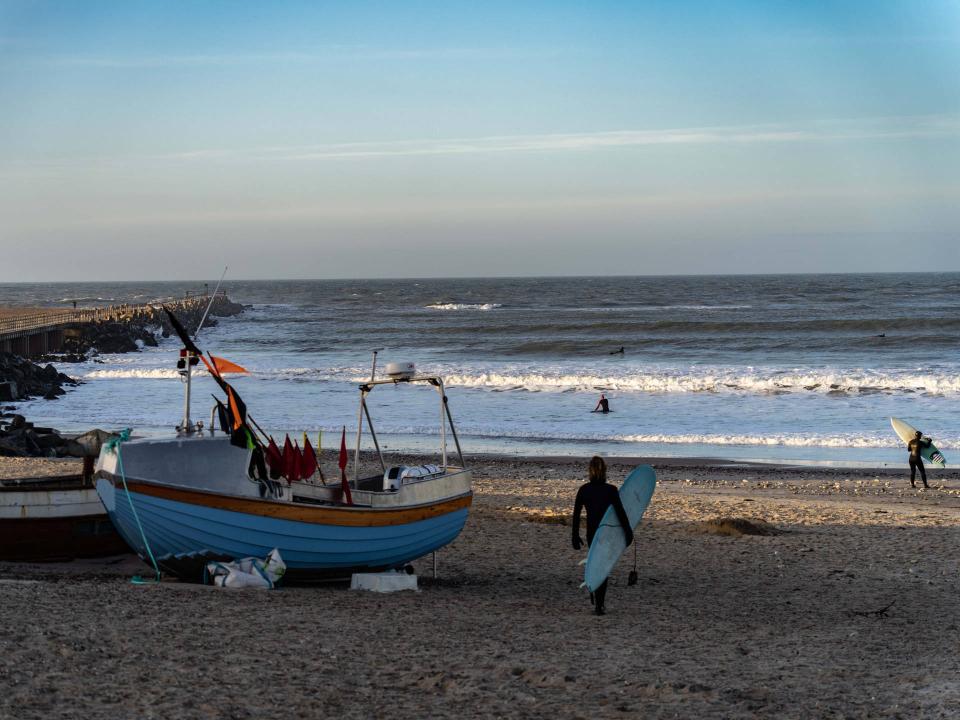 Verwittertes Fischerboot wartet am sandigen Strand während ein Surfer mit Brett zu den Wellen geht____