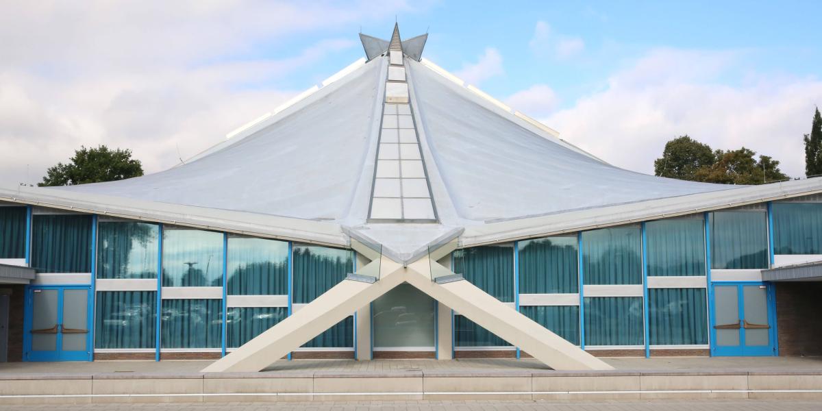 Modernes Gebäude mit geschwungenem Dach und großen Glasfassaden, kontrastiert von klarer blauer und weißer Architektur.