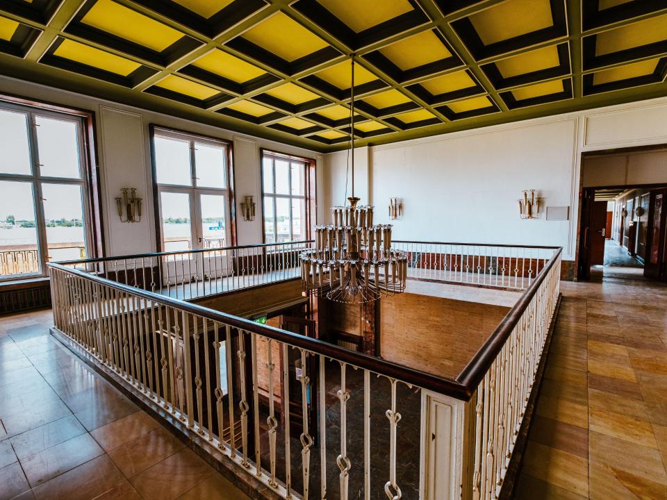 Innenansicht eines Gebäudes mit gelber Kassettendecke, großen Fenstern, Treppengeländer und Kronleuchter.____