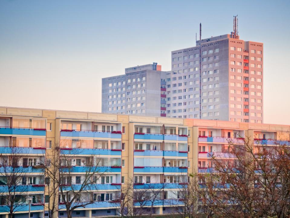 Wohnkomplex mit bunten Balkonen und einem dahinterliegenden Hochhaus, beleuchtet von weichem Abendlicht.____