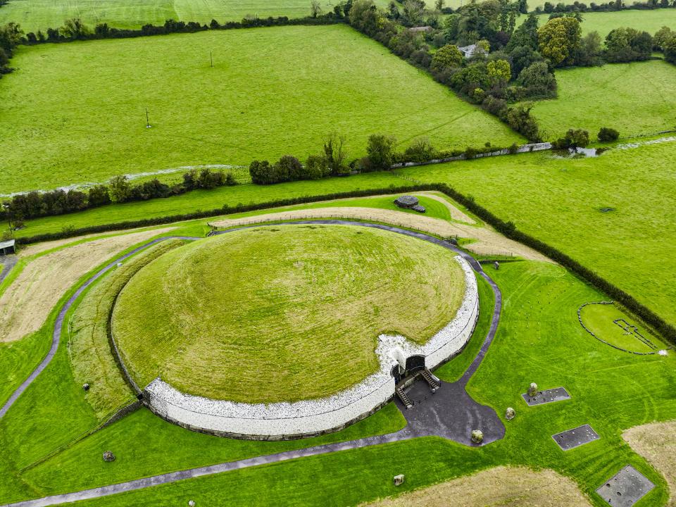 Großes grasbedecktes Hügelgrab mit weißem Steinsockel und dunklem Zugangspfad in ländlicher Umgebung.____