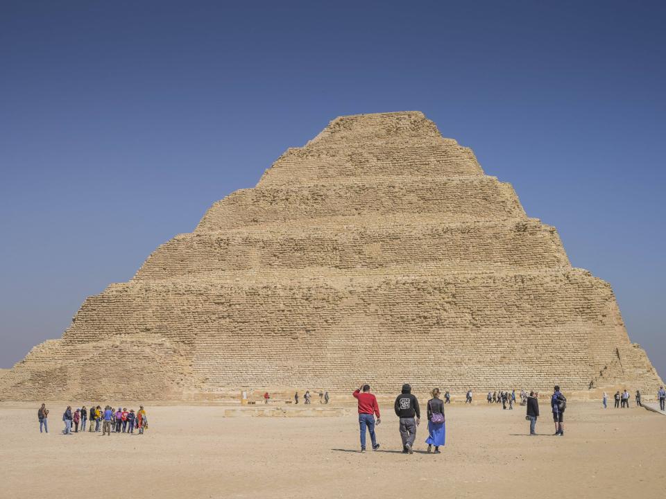 Die Stufenpyramide ragt aus der Wüste empor, umgeben von Besuchern auf hellem Sandboden.____