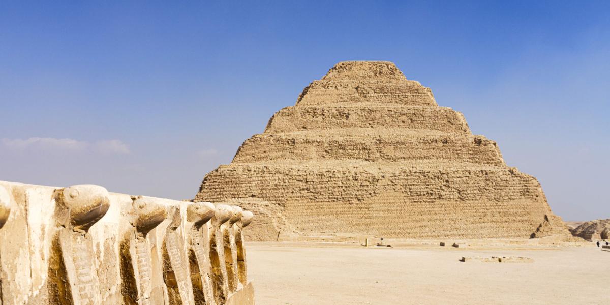 Antike Stufenpyramide aus Kalkstein mit monumentalem Sockel im Vordergrund, gelegen in weiter Wüstenlandschaft.