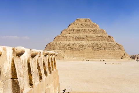 Antike Stufenpyramide aus Kalkstein mit monumentalem Sockel im Vordergrund, gelegen in weiter Wüstenlandschaft.