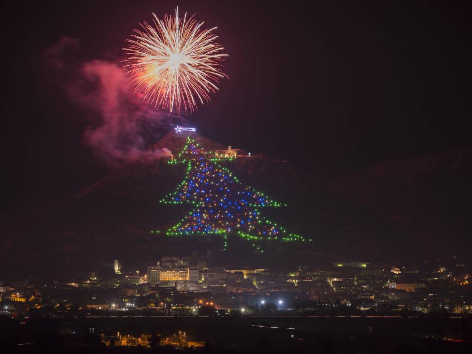 Lichterbaum am Hang in Gubbio, Italien, mit bunten Lichtern und Feuerwerk am Nachthimmel.____