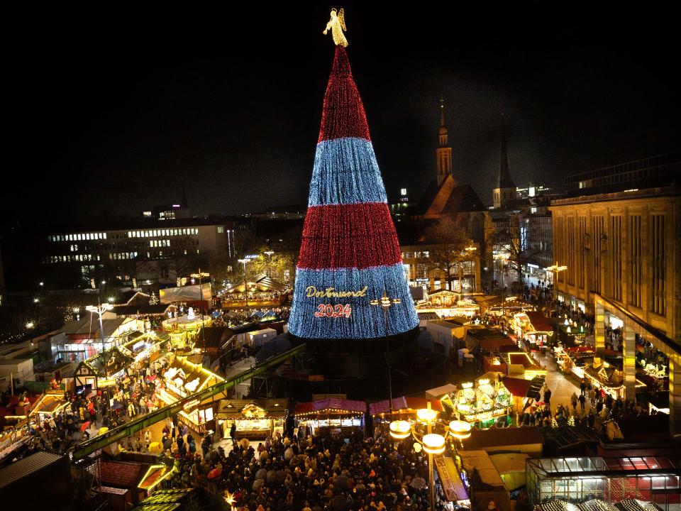 Weihnachtsbaum in Dortmund bei Nacht, beleuchtet mit rot-blauen Lichtern, umgeben von Weihnachtsmarktständen.____