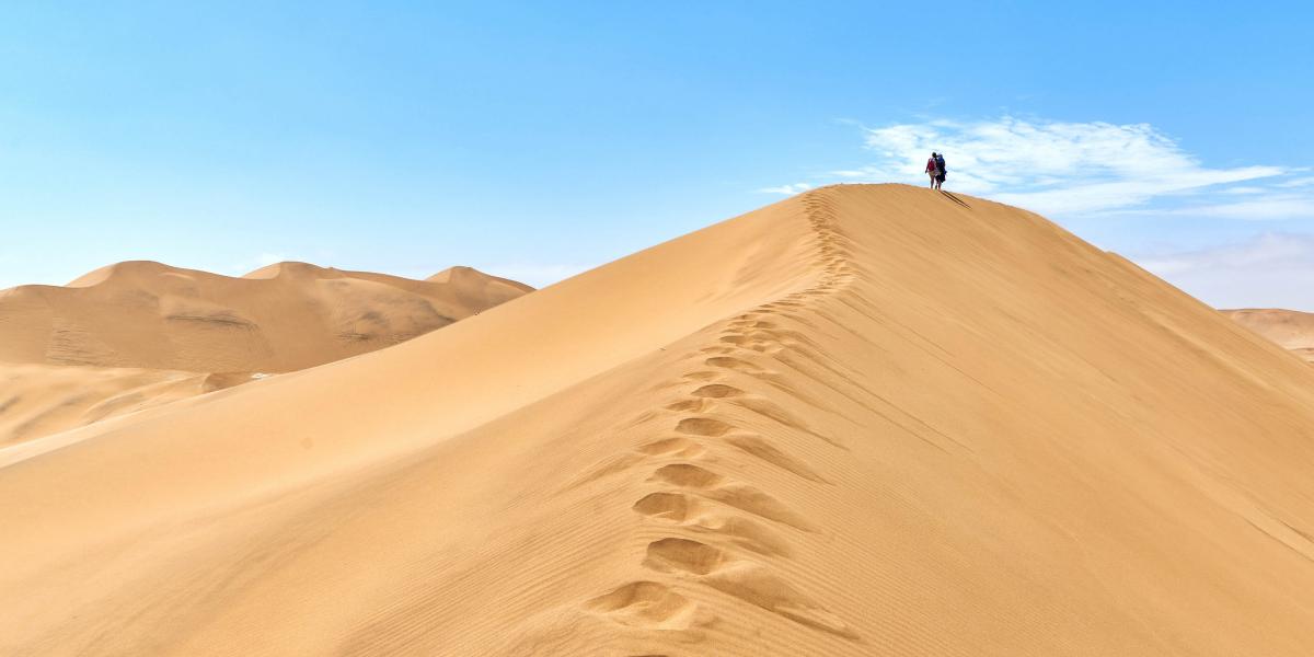 Fußspuren führen auf eine hohe Sanddüne, einsamer Wanderer unter klarem Himmel.