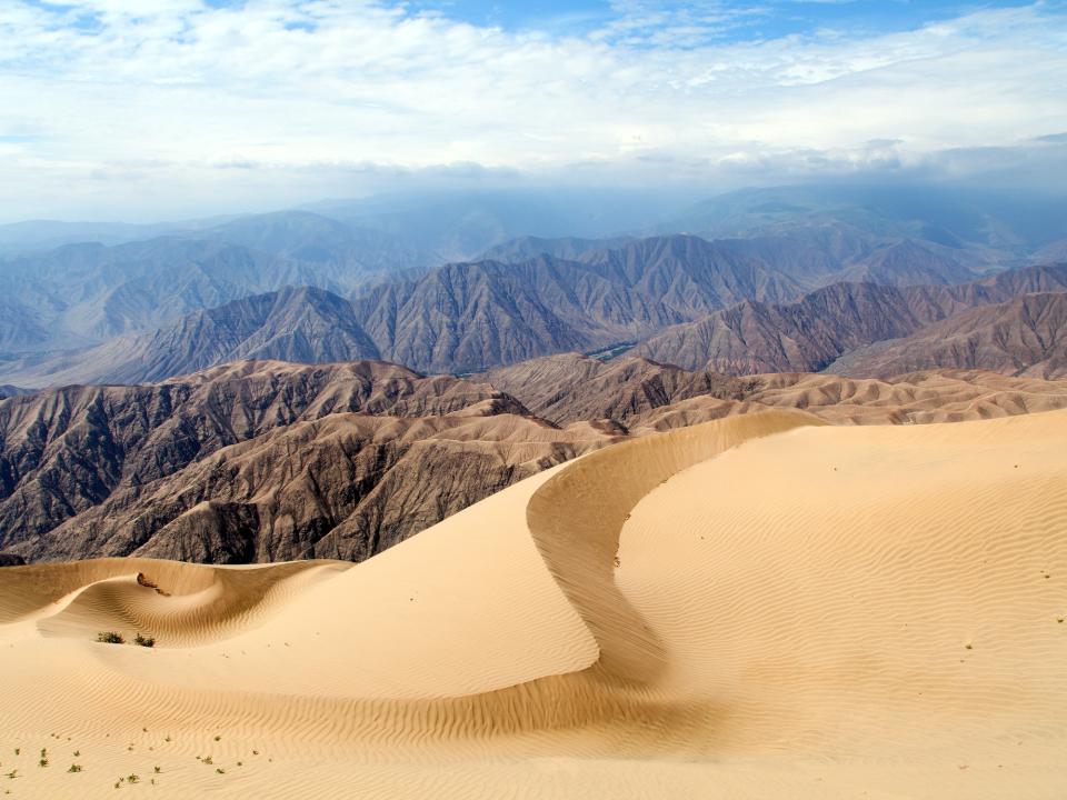 Sanddünen im Vordergrund mit Bergen im Hintergrund und teils bewölktem Himmel.____