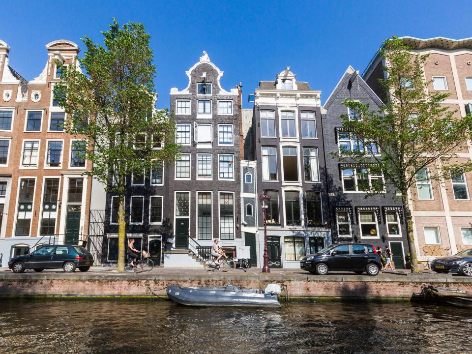 Amsterdamer Grachtenhäuser mit verzierten Fassaden und Bäumen, ein Boot auf dem Kanal bei klarem Himmel.____