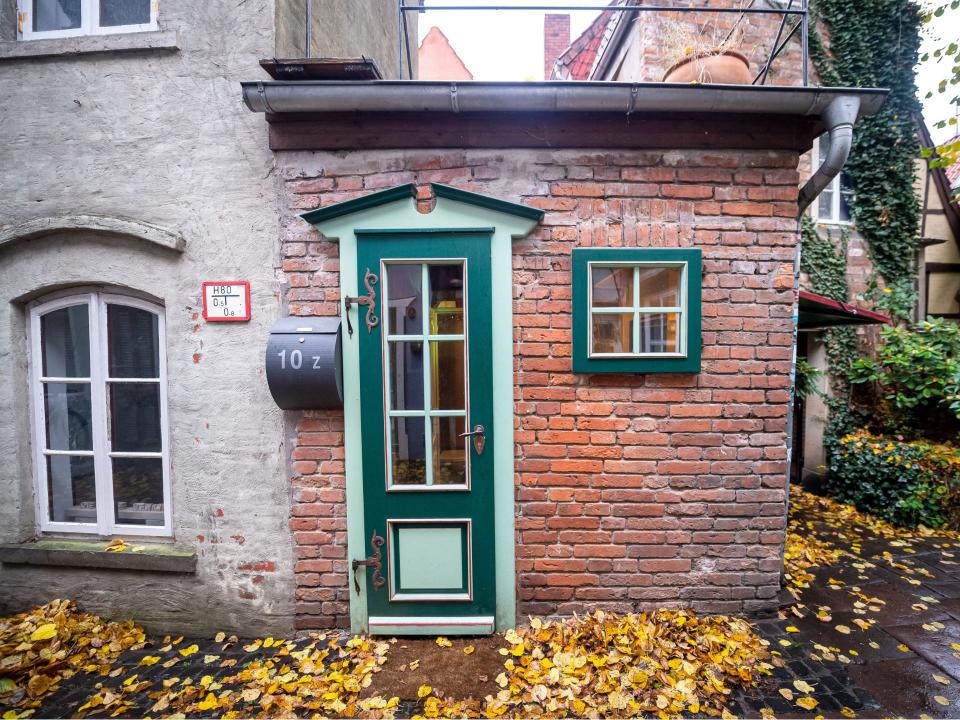 Kleines Backsteinhaus mit grüner Tür und Fenstern, Herbstlaub bedeckt den Boden, malerisches Detail.____