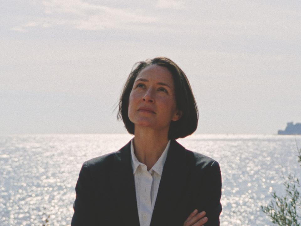 Frau im schwarzen Blazer blickt nachdenklich aufs Meer, Sonnenlicht reflektiert auf dem Wasser.____