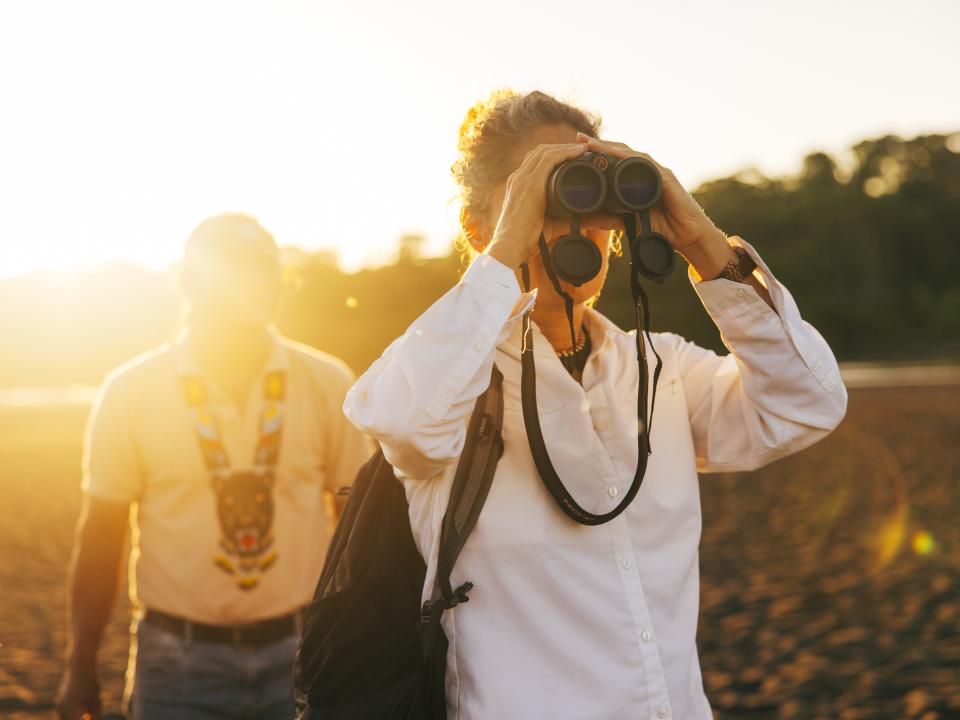 Frau mit Fernglas beobachtet bei Sonnenuntergang die Natur, begleitet von einem weiteren Mann im Hintergrund.____