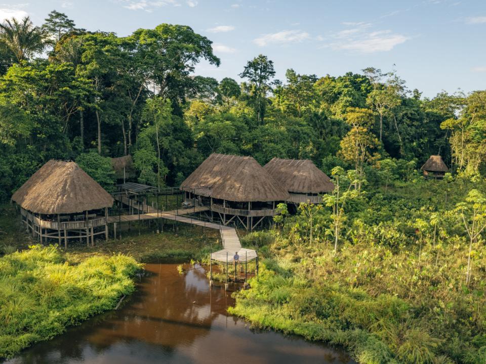 Ökolodge mit strohgedeckten Hütten, Holzstegen und üppigem tropischen Wald, nahe einem Flusslauf.____