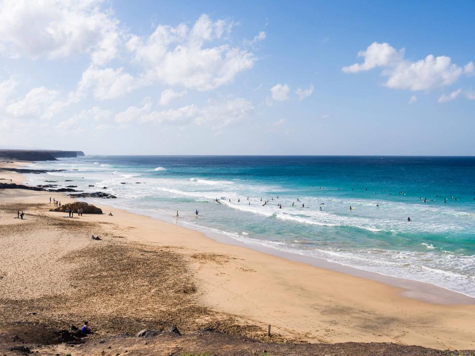 Mehrere Surfer paddeln auf ruhigen Wellen vor einem weiten Sandstrand unter blauem Himmel____