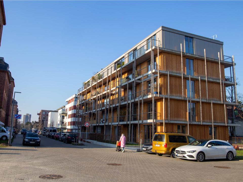 Modernes Wohngebäude mit Holzfassade, geparkte Autos und Gehweg bei sonnigem Wetter, urbanes Wohngebiet.____